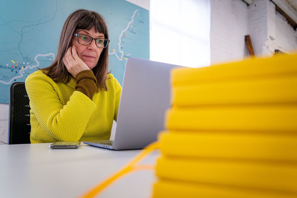 Jackie Jones wearing yellow at her desk in the Super Slow Way Offices with yellow notebooks in the foreground. Image by Rachel Ovenden.