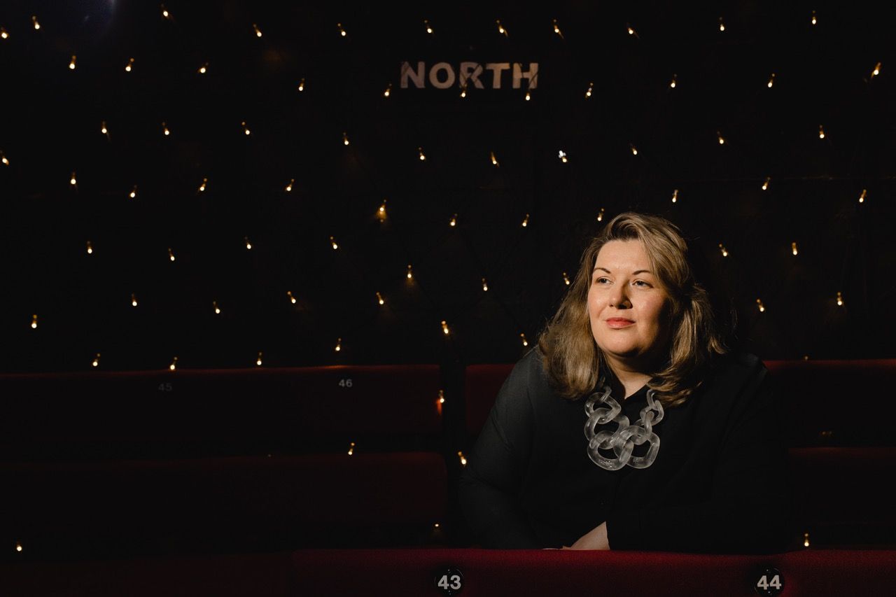 Karen O'Neill seated in The Dukes Theatre with North Signage in the background. Image by Christina Davies.