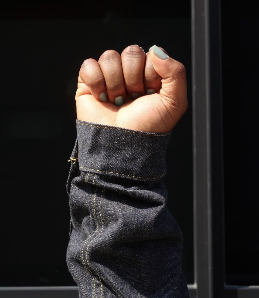 Black women's fist and denim sleeve against black background, part of Tiwirayi Ndoro's Woke Denim Project