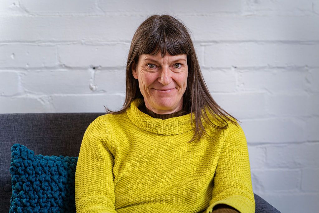 Jackie Jones wearing a yellow sweater seated on a grey couch against a white brick wall. Image by Rachel Ovenden.