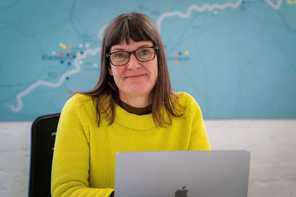 Jackie Jones in a yellow sweater in front of her computer with backdrop of white wall and blue map. Image by Rachel Ovenden.